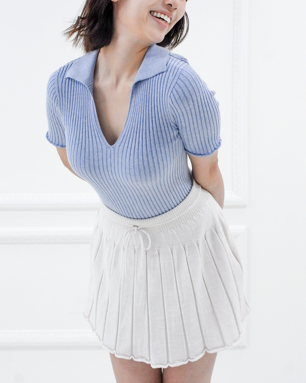 Close three-quarter view of the Corinne ribbed cotton knit polo in Cielo blue, styled with a white pleated skirt