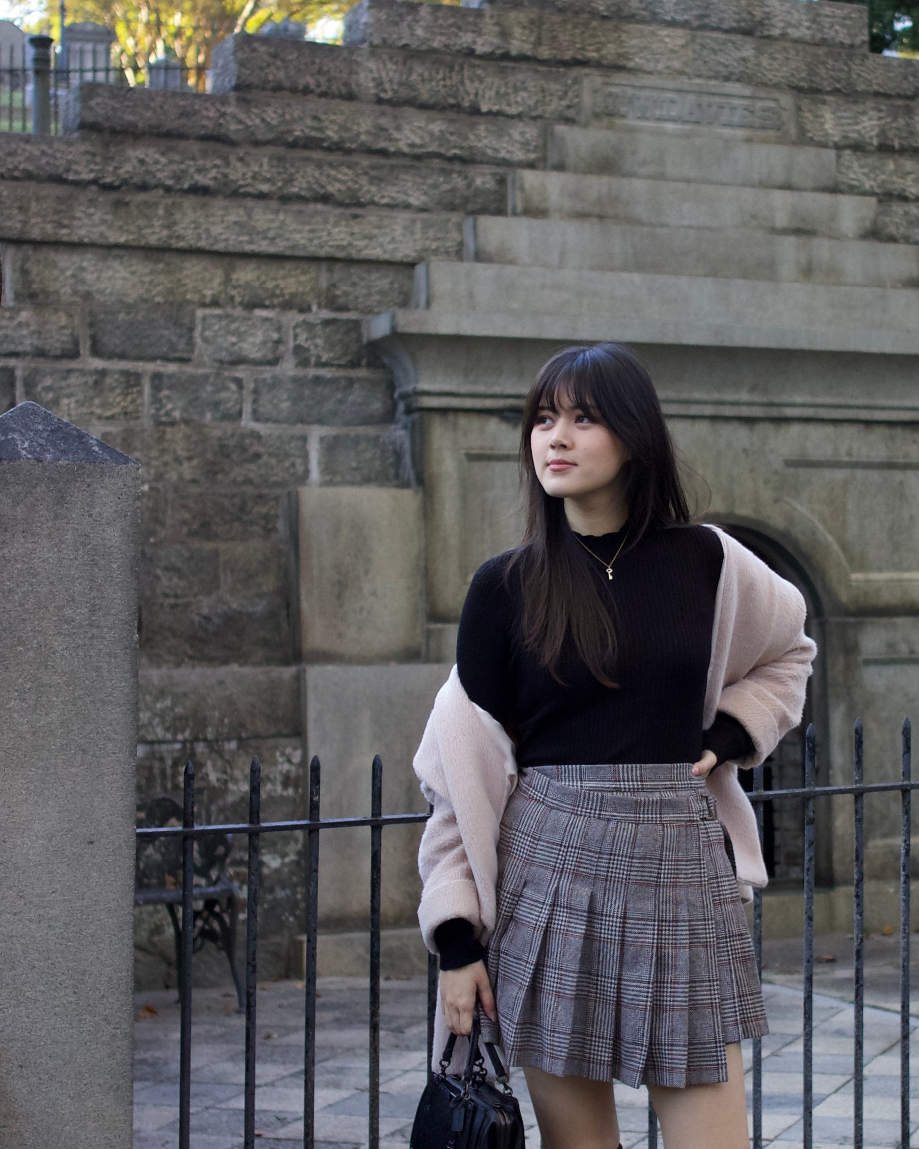 Woman standing in front of a stone wall wearing a black top, plaid skirt, and beige coat.