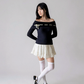 Woman wearing a dark blue off-shoulder top and white skirt with white stockings and black shoes on a plain background