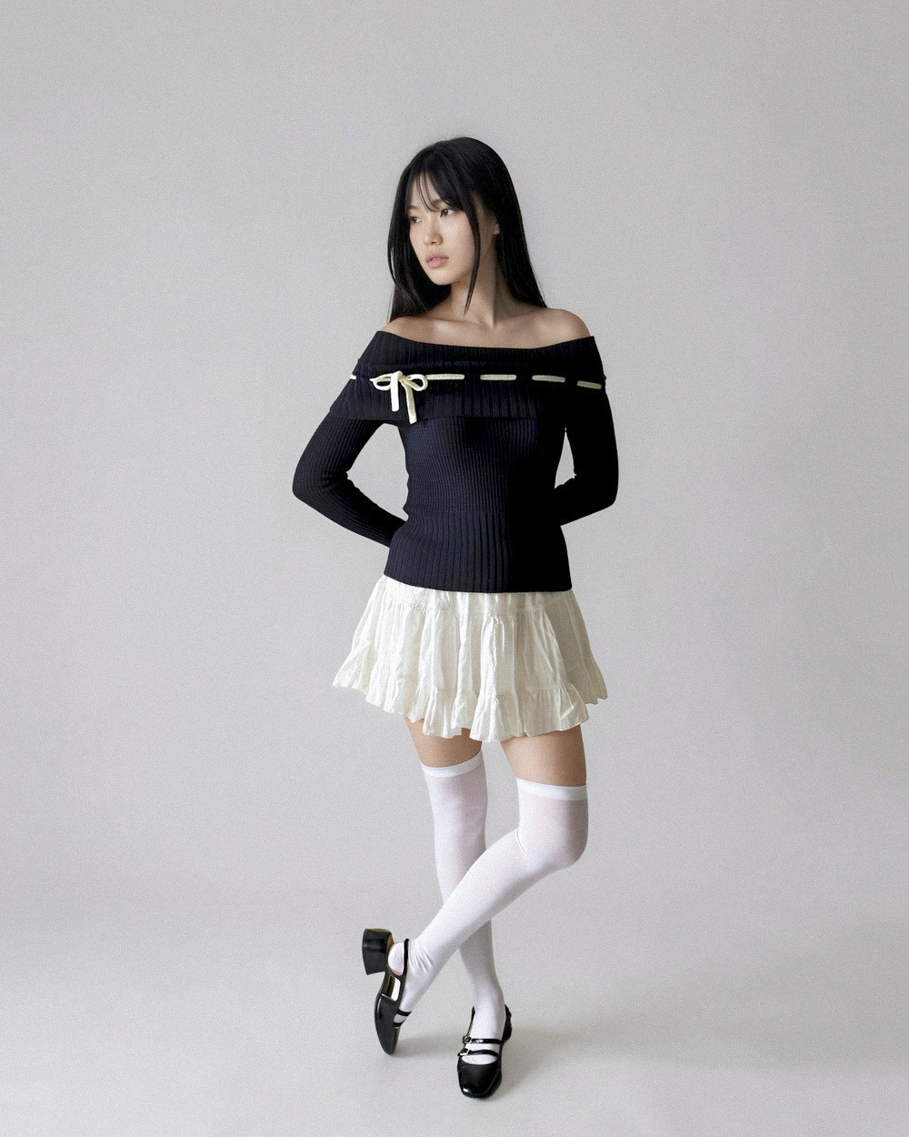 Woman wearing a dark blue off-shoulder top and white skirt with white stockings and black shoes on a plain background