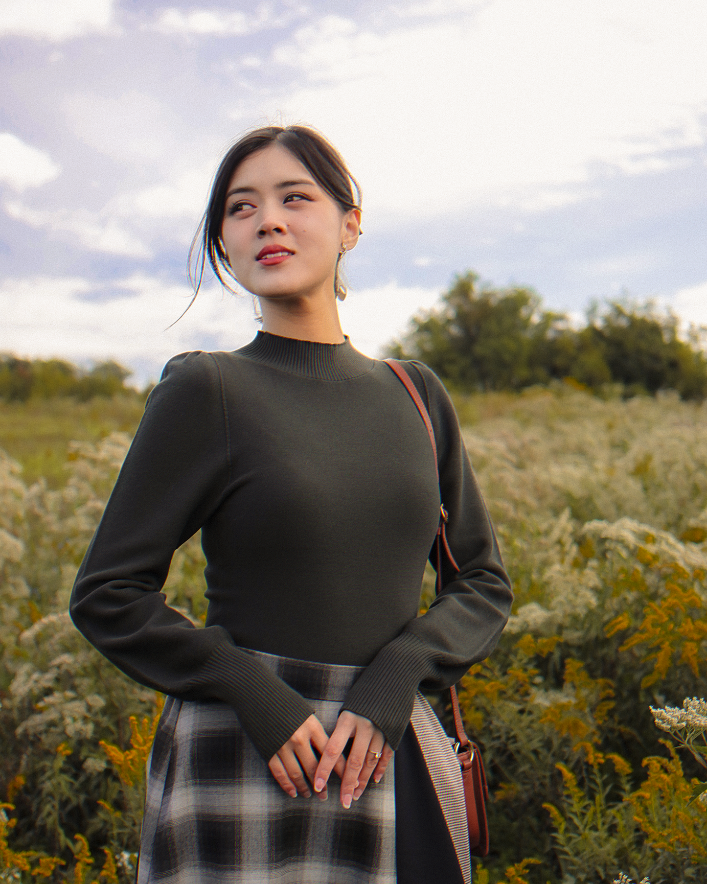 A woman posing in the Meryl top in the color Pine, a muted dark green color. There is a meadow in the background.