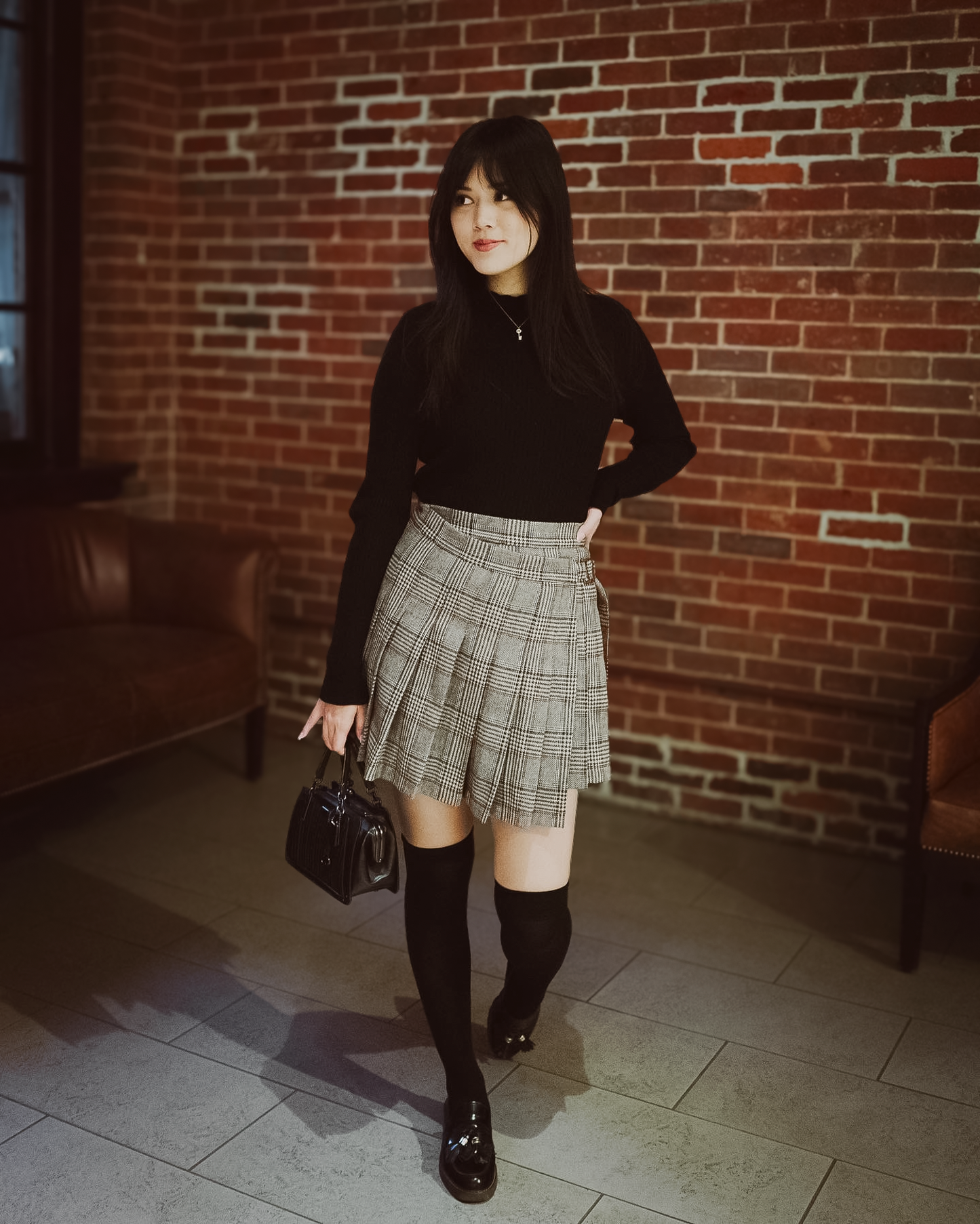 Woman in black top and plaid skirt standing against a brick wall.