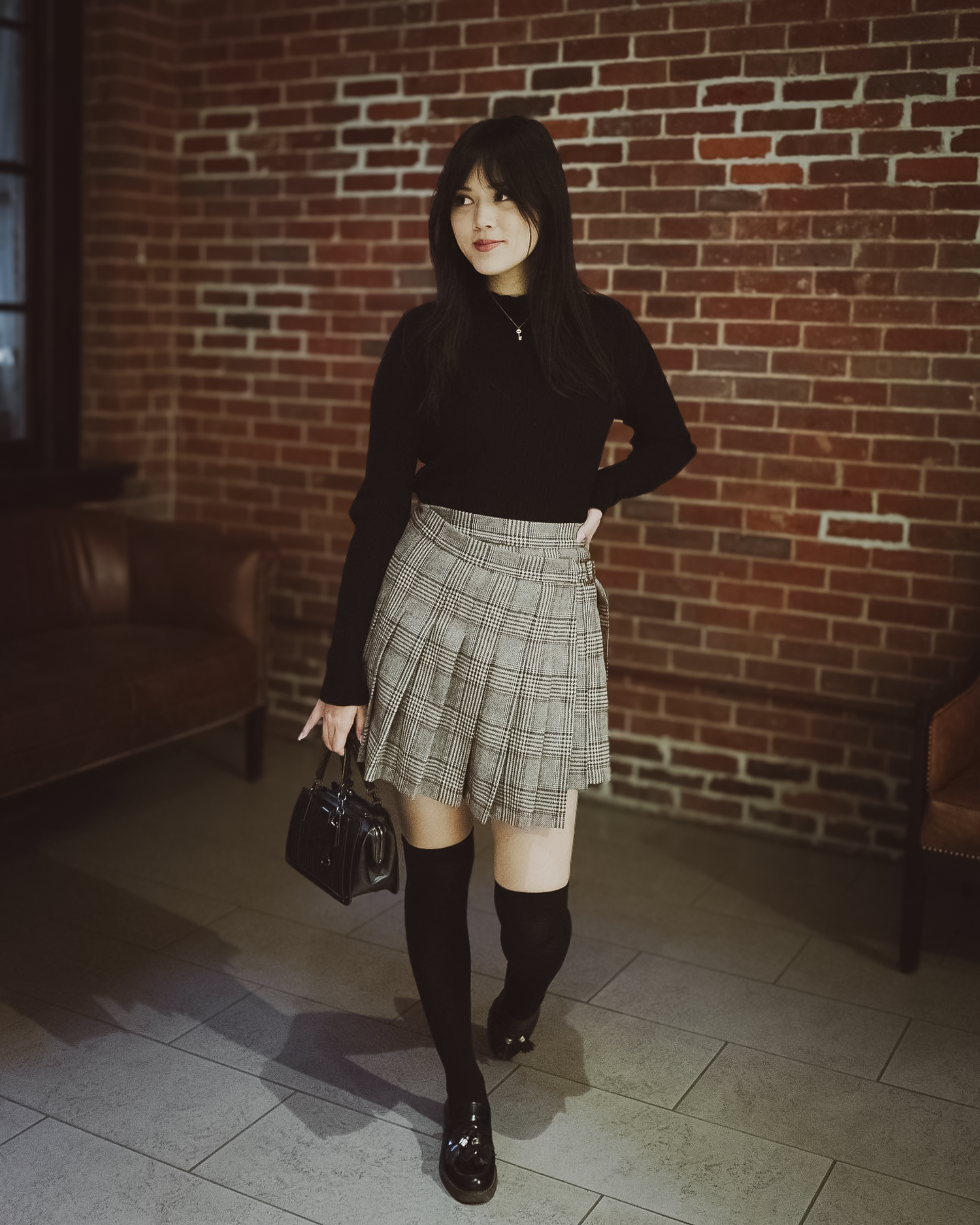 Woman in black top and plaid skirt standing against a brick wall.